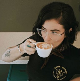 A woman in eyeglasses holds a foamed coffee drink to her mouth, wearing a Dark Horse Coffee Roasters tee shirt