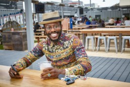 A smiling man wearing a hat and a vibrantly patterned shirt holds a cup of tea in one hand and his phone in the other.