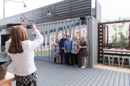 A woman takes a photo on her phone of a group of people in front of a steel container